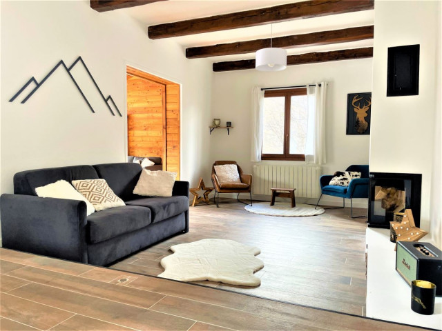 Grand séjour lumineux avec cheminée, canapé, fauteuils, ameublement bois, poutres apparentes, parquet au sol, fenêtre