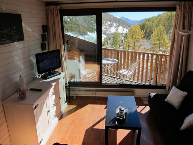 Bright studio living room with sofa, dining table, TV, French doors opening onto a wooden balcony with mountain view